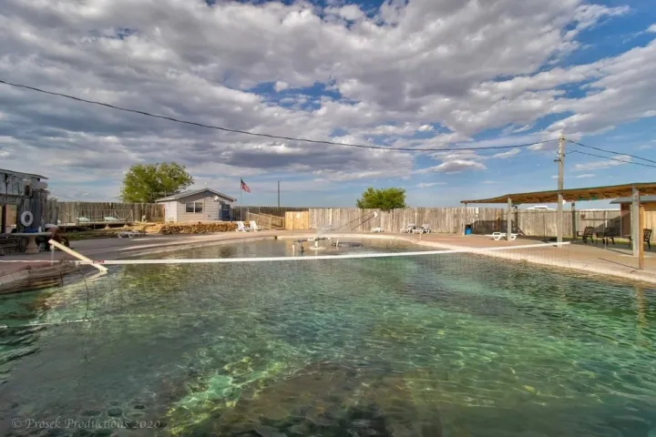 Outdoor swimming pool with cloudy sky, surrounded by fences and a small building.